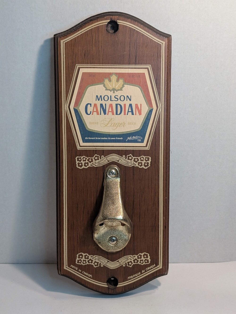 A vintage Molson Canadian branded bottle opener mounted on a wooden wall plaque.